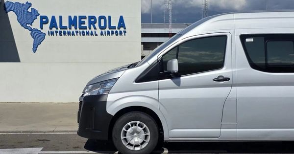 White passenger van parked near the Palmerola International Airport sign, with a blue map silhouette in the logo visible on a light-colored wall.